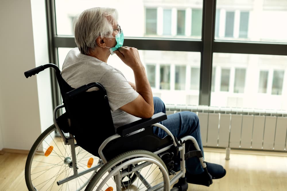 Mature man in wheelchair feeling lonely and looking through the window while being in home quarantine due to coronavirus epidemic. 